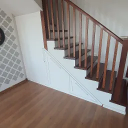 Drewniane schody z brązowymi stopniami i białą balustradą, zintegrowane z białą szafą wnękową pod schodami, na ścianie wzór geometryczny i okrągłe lustro.