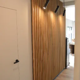 Drewniana ścianka działowa z pionowych listew oświetlona trzema czarnymi reflektorami, widok z korytarza obok białych drzwi z czarną klamką.