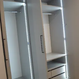 Otwarta szara szafa z oświetleniem LED, widoczne półki, drążek na ubrania i dwie szuflady, minimalistyczny design.