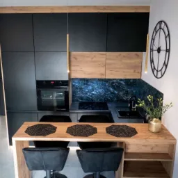 Nowoczesna kuchnia z czarnymi szafkami, drewnianymi akcentami i marmurowym backsplashiem, oświetlona złotymi lampami wiszącymi, z drewnianym blatem barowym i czarnymi hokerami.