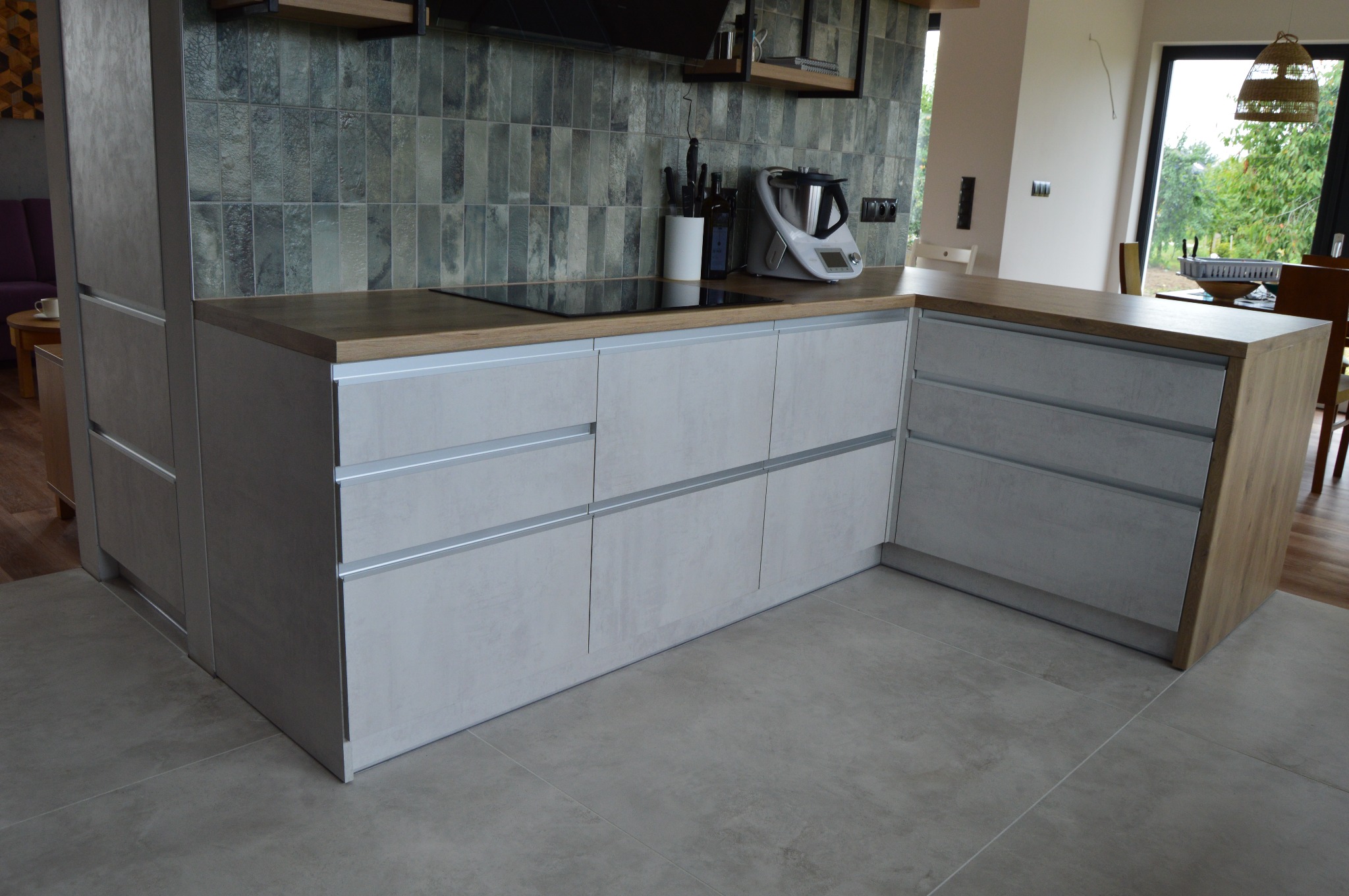 Szara, nowoczesna kuchnia z drewnianym blatem, płytą indukcyjną i sprzętem kuchennym na blacie w tle okno z widokiem na zieleń.