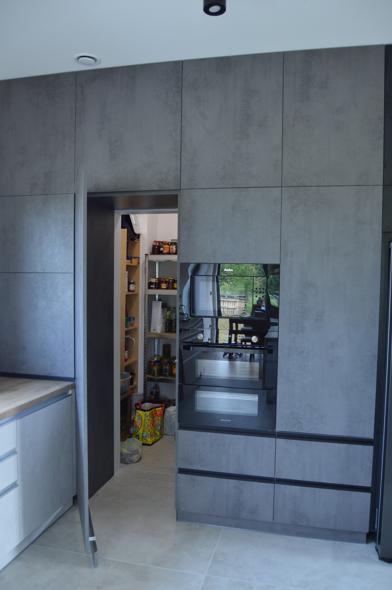 Szara zabudowa kuchenna z piekarnikiem i spiżarnią ukrytą za drzwiami, widoczne półki spiżarni wypełnione produktami spożywczymi.