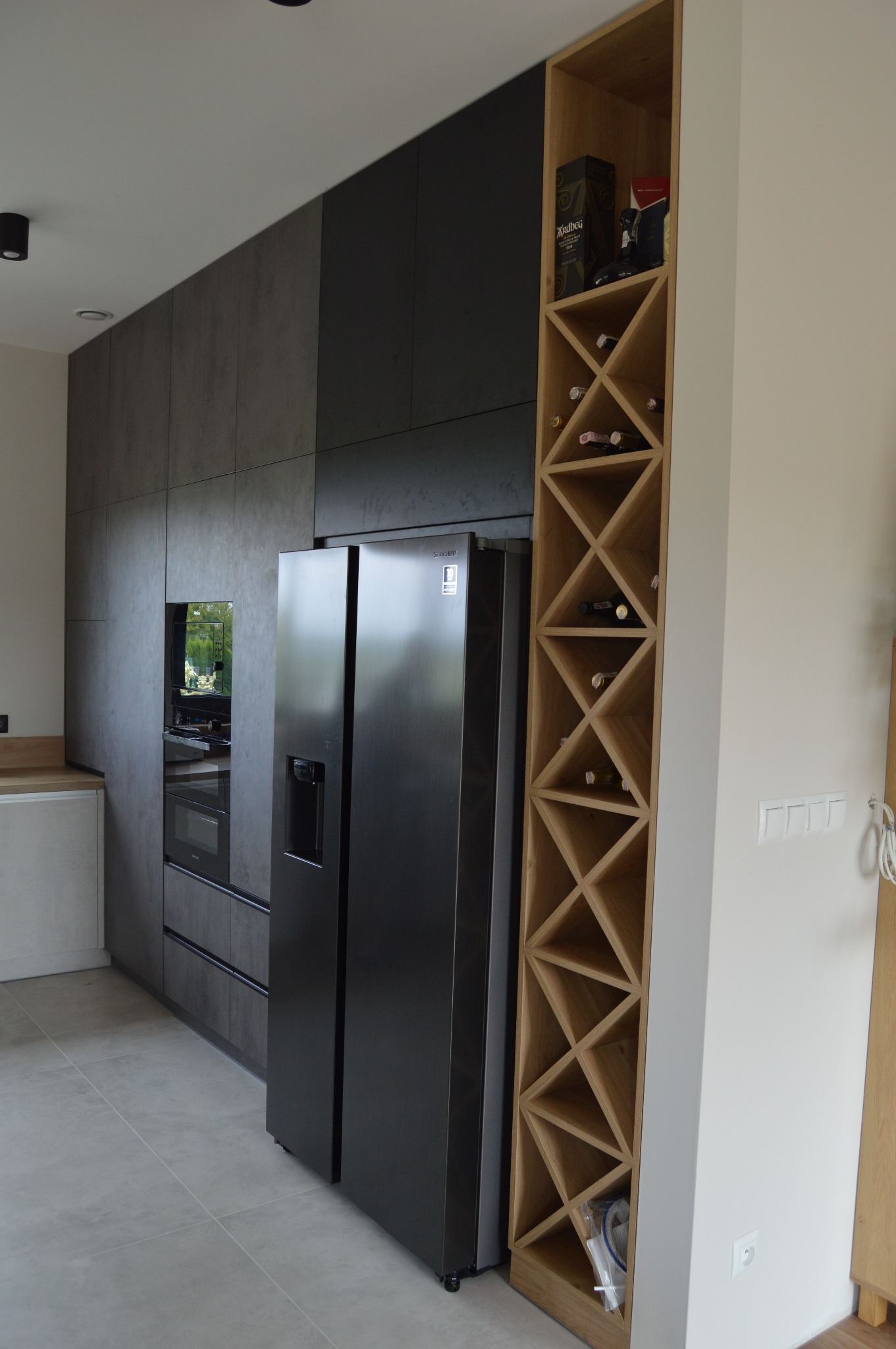Nowoczesna kuchnia z szarymi frontami, zabudowaną lodówką i drewnianym regałem na wino z trójkątnymi przegródkami.