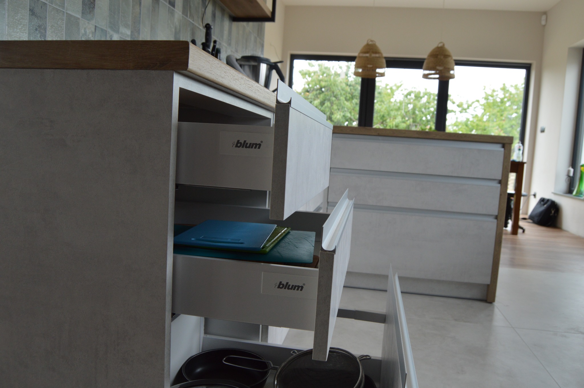 Szare szafki kuchenne z otwartymi szufladami, w których widoczne są deski do krojenia i naczynia. Widoczne logo Blum na szufladach.