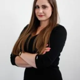 Portret kobiety w czarnej sukience z założonymi rękami na tle białej ściany, z widocznym zegarkiem na lewym nadgarstku.