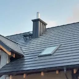 Nowy dach z ciemnej dachówki ceramicznej, z oknem dachowym i metalowym kominem, w trakcie budowy domu.