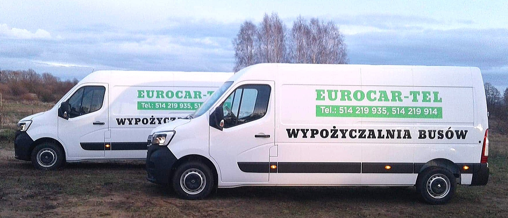 Dwa białe busy z zielonym logo i napisem 'WYPOŻYCZALNIA BUSÓW' na tle pochmurnego nieba i drzew bez liści, stojące na ziemi.