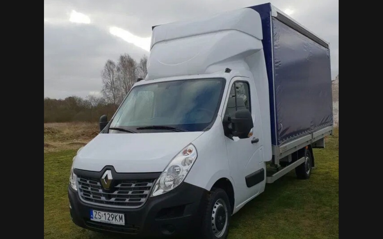 Biały samochód dostawczy Renault Master z plandeką w kolorze granatowym, zaparkowany na trawiastym terenie z widocznym tłem drzew.