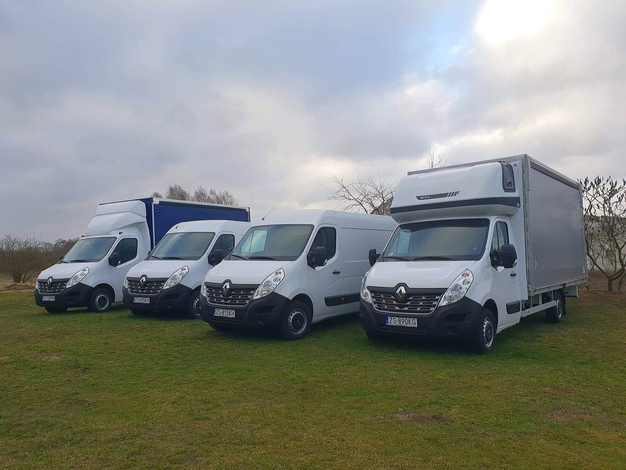 Cztery białe busy Renault Master z różnymi zabudowami kontenerowymi stojące w rzędzie na trawiastym terenie pod pochmurnym niebem.