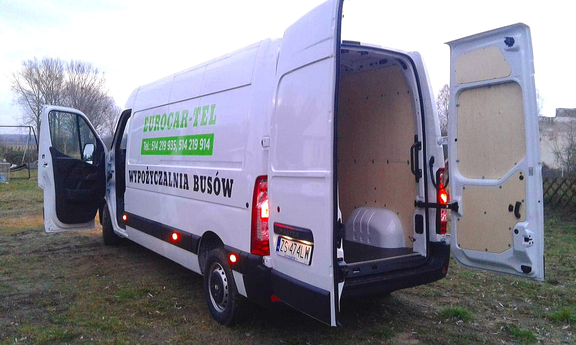 Biały bus z otwartymi drzwiami bocznymi i tylnymi, widoczny napis 'WYPOŻYCZALNIA BUSÓW' oraz numer telefonu, zaparkowany na trawiastym terenie.
