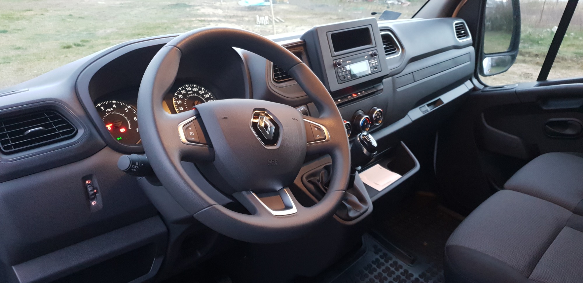 Wnętrze kabiny kierowcy Renault Master: kierownica z logo, deska rozdzielcza z radiem i klimatyzacją, widoczne wskaźniki i dźwignia zmiany biegów, szare fotele.