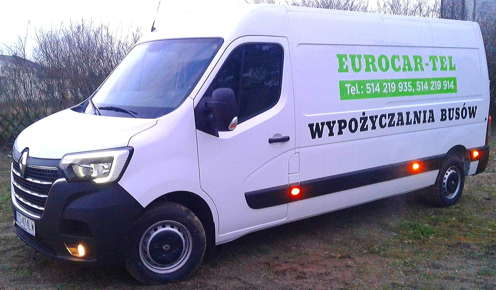 Biały bus Renault Master z napisem 'Wypożyczalnia Busów' i logo firmy Eurocar-Tel z numerami telefonów, zaparkowany na trawiastym terenie