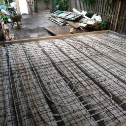 AlpinSpec Bud Grajewo - Przygotowanie podłoża pod wylewkę z ogrzewaniem podłogowym; widoczne rury grzewcze zamocowane do siatki zbrojeniowej, w tle narzędzia i materiały budowlane.