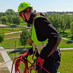 AlpinSpec Bud Grajewo - Pracownik w kasku i uprzęży linowej, przygotowuje się do pracy na wysokości, z widokiem na park miejski w tle.