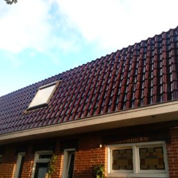 AlpinSpec Bud Grajewo - Czerwono-czarny dach z dachówki ceramicznej z oknem dachowym, widok z dołu na tle błękitnego nieba, ściana budynku z czerwonej cegły.