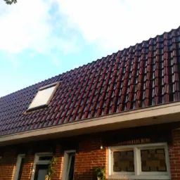 Czerwono-czarny dach z dachówki ceramicznej z oknem dachowym, widok z dołu na tle błękitnego nieba, ściana budynku z czerwonej cegły.