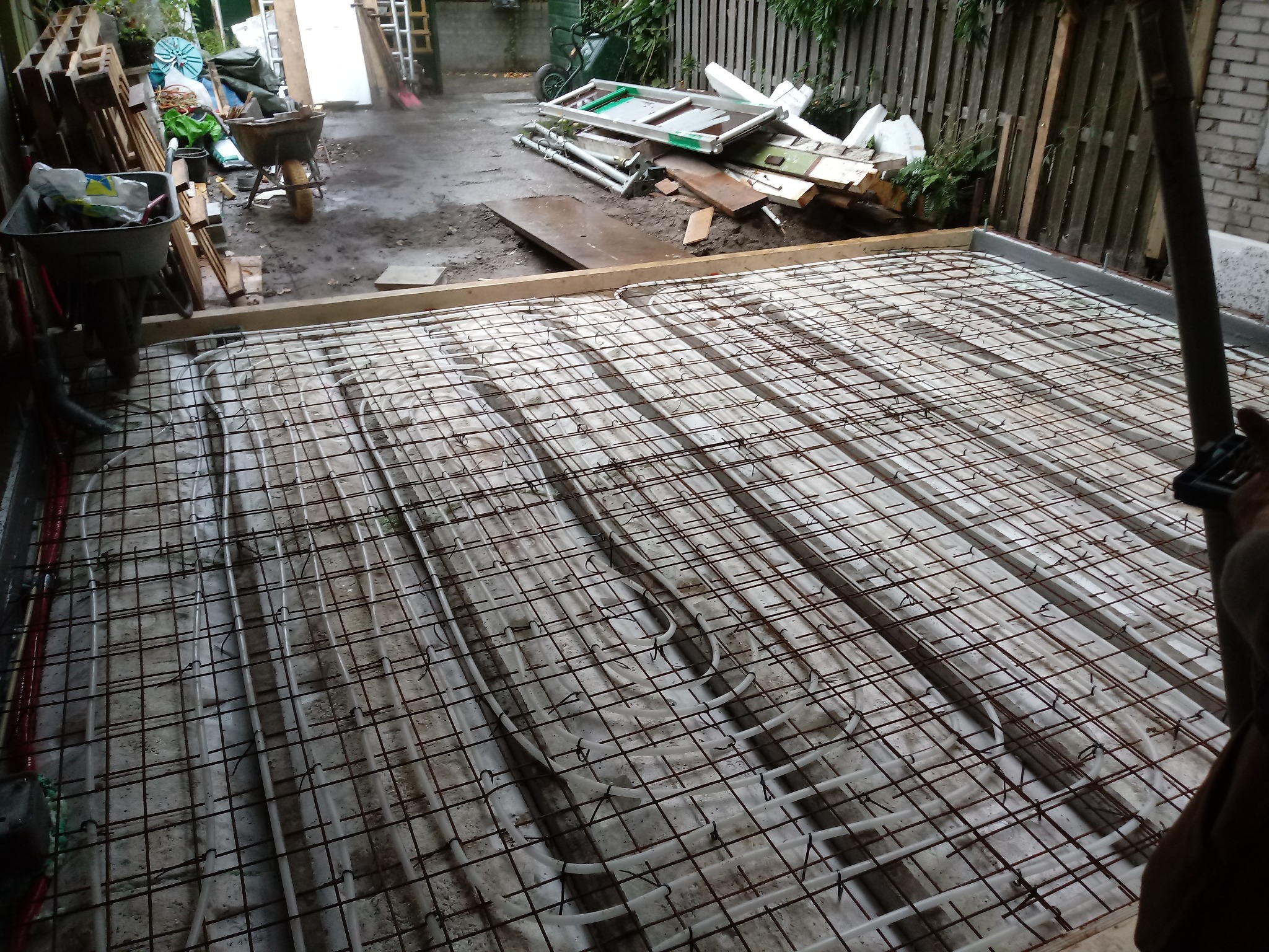 Przygotowanie podłoża pod wylewkę z ogrzewaniem podłogowym; widoczne rury grzewcze zamocowane do siatki zbrojeniowej, w tle narzędzia i materiały budowlane.