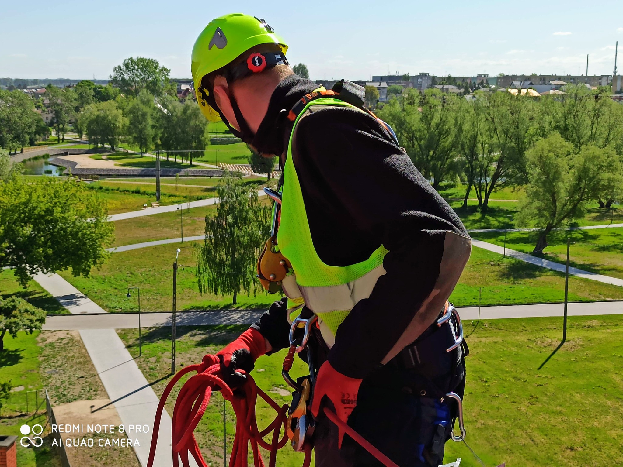 Pracownik w kasku i uprzęży linowej, przygotowuje się do pracy na wysokości, z widokiem na park miejski w tle.