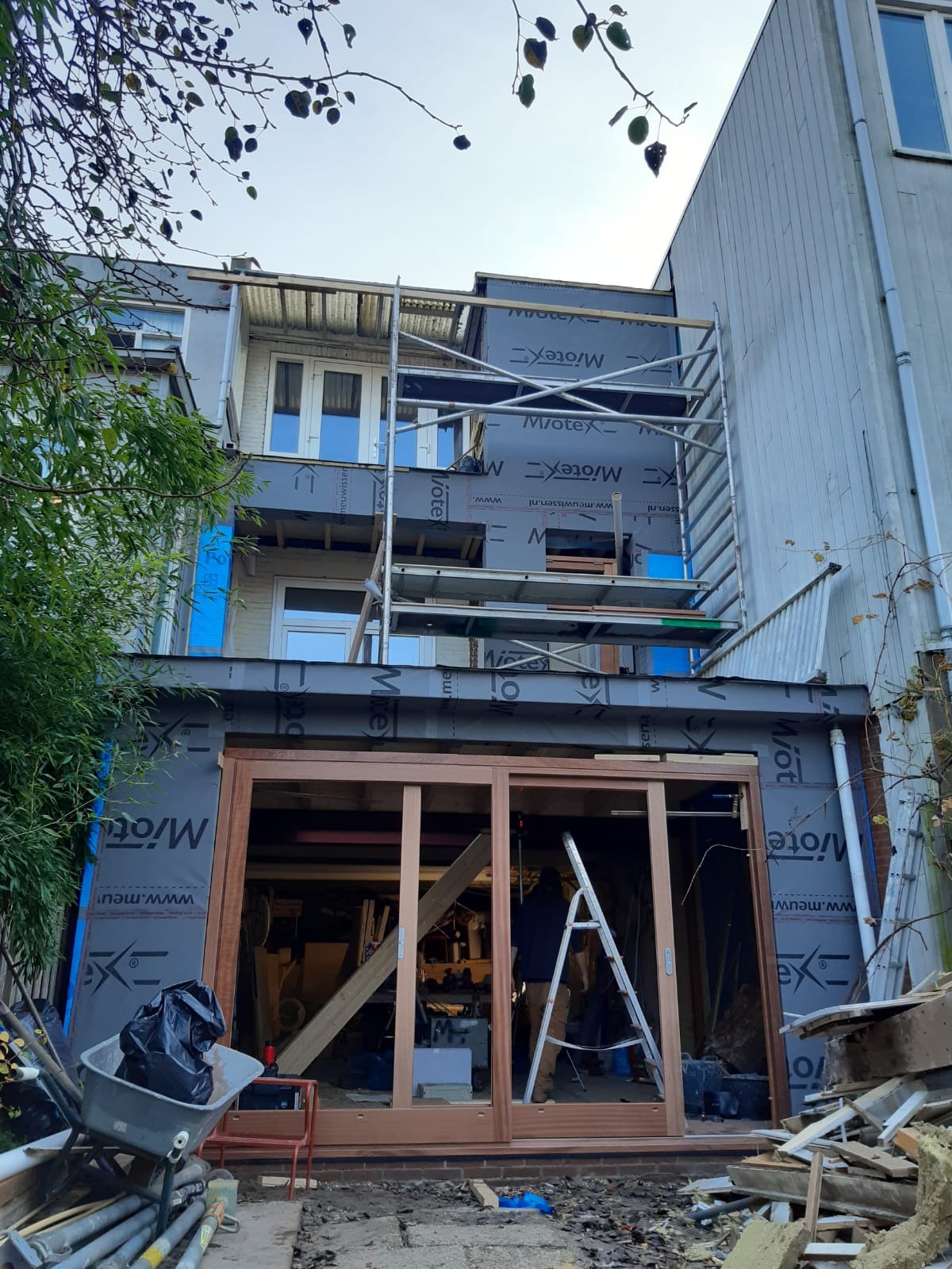 Budynek w trakcie remontu, widoczne rusztowania, materiały izolacyjne z nadrukiem, częściowo zamontowane drewniane ramy okienne, drabina i gruz na pierwszym planie.