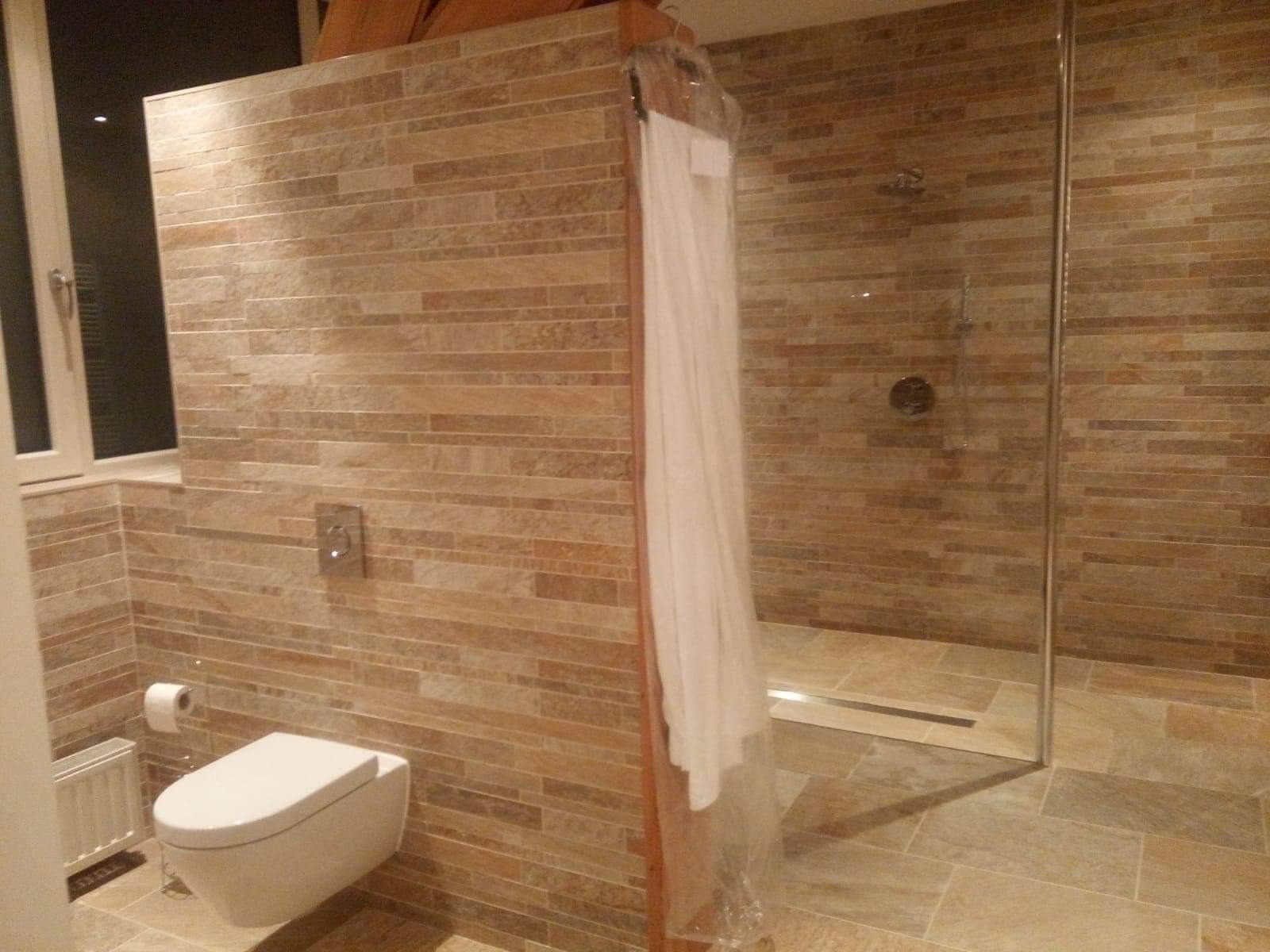 Nowoczesna łazienka z beżowymi płytkami imitującymi kamień, wiszącą toaletą i kabiną prysznicową typu walk-in, oddzieloną od reszty pomieszczenia ścianką. Biały szlafrok w folii ochronnej...