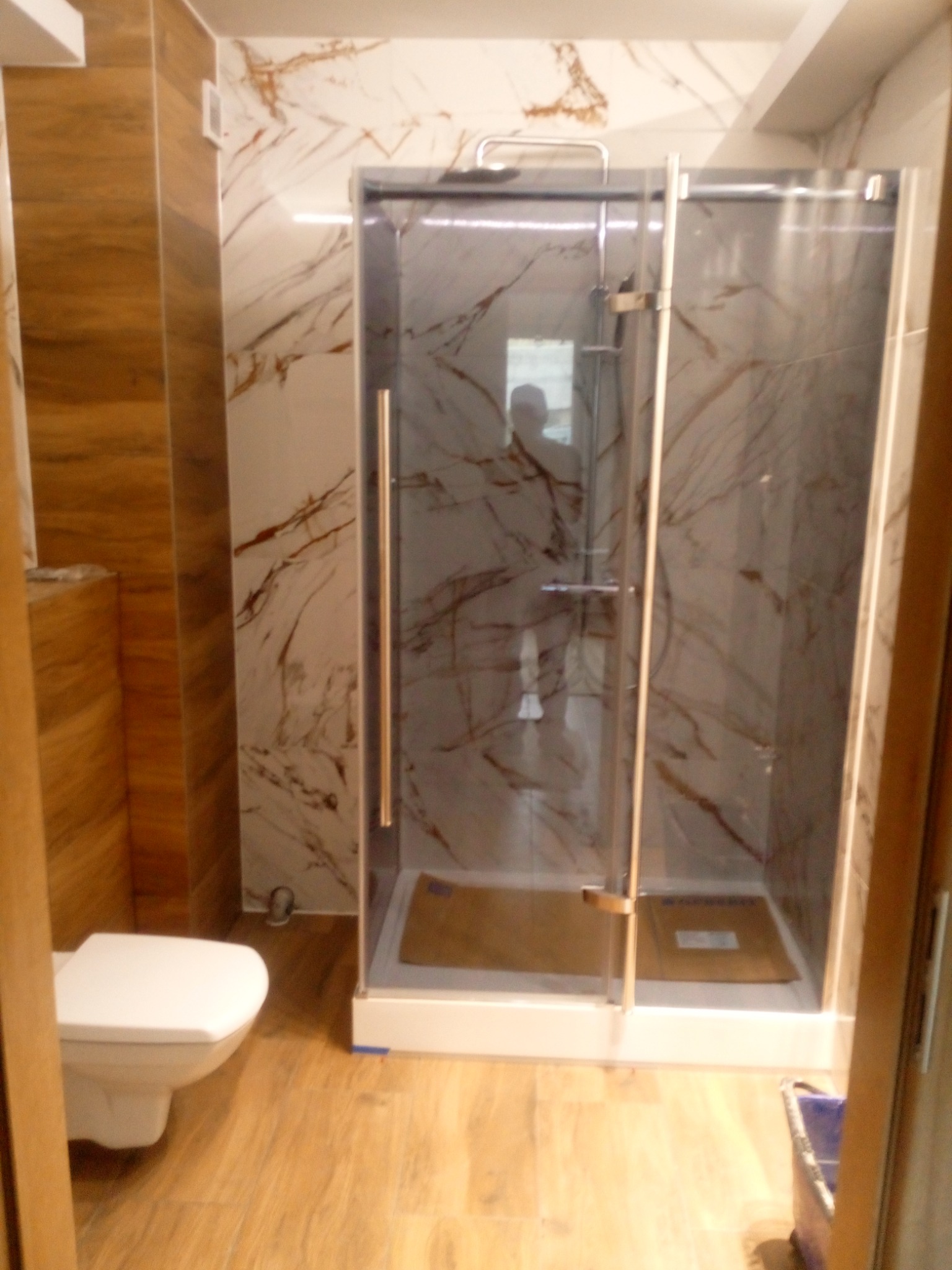 Nowoczesna kabina prysznicowa z marmurowymi płytkami w odcieniach szarości i brązu, widoczna toaleta i drewnopodobne płytki podłogowe.