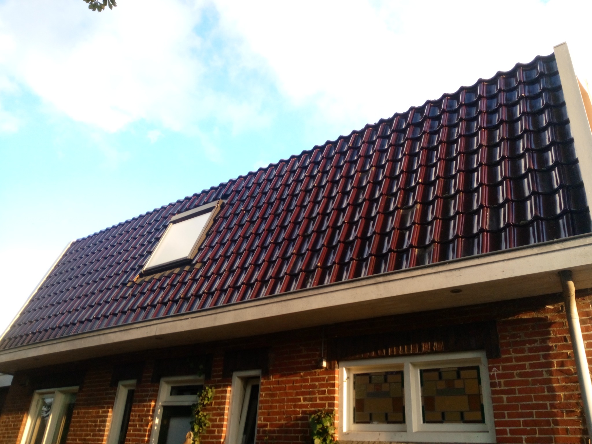 Czerwono-czarny dach z dachówki ceramicznej z oknem dachowym, widok z dołu na tle błękitnego nieba, ściana budynku z czerwonej cegły.