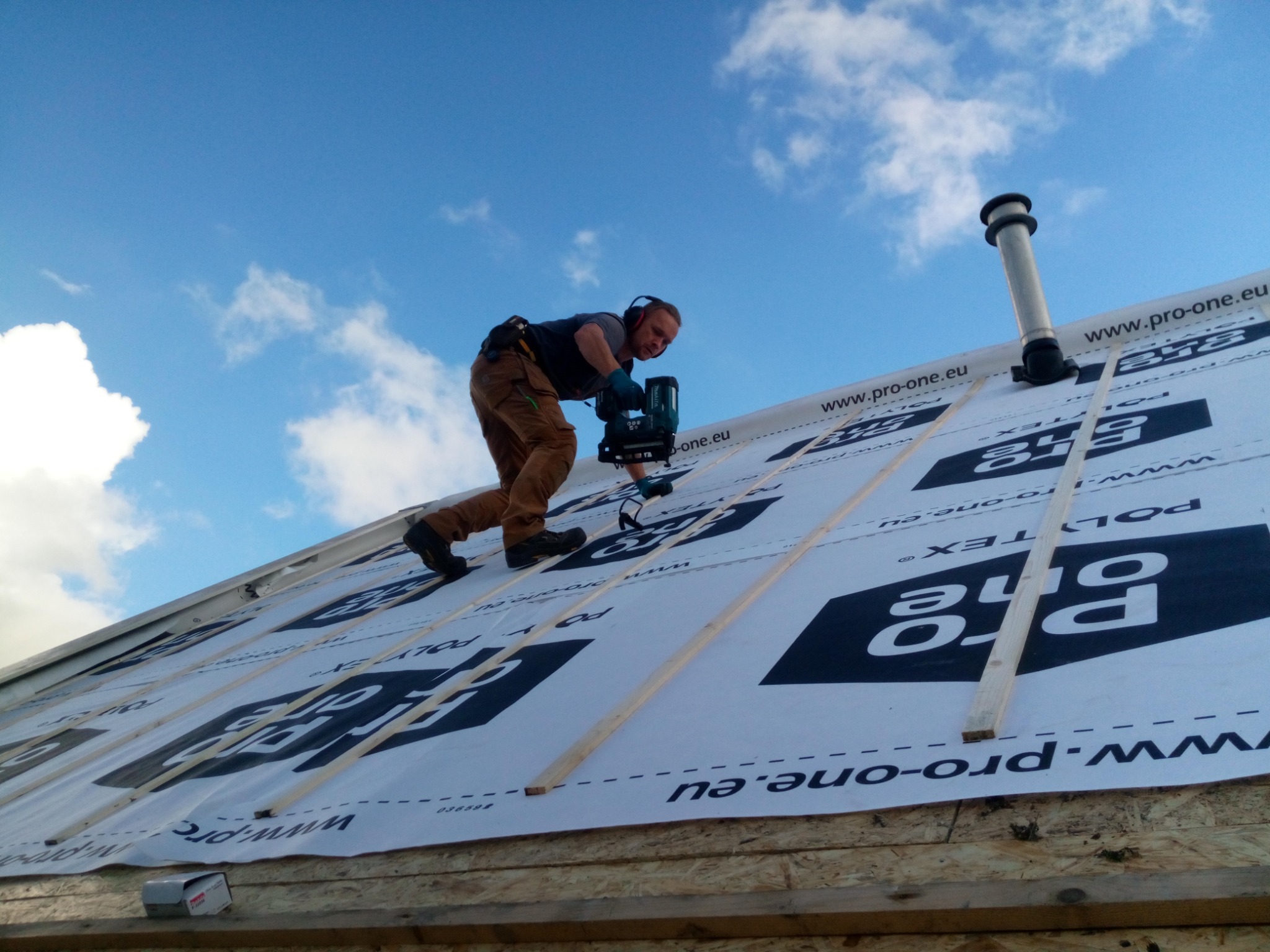 Pracownik w kasku i słuchawkach ochronnych, pochylony na dachu pokrytym membraną z widocznym nadrukiem 'Pro-one', używa narzędzia do mocowania łat dachowych, na tle błękitnego nieba z chmurami.
