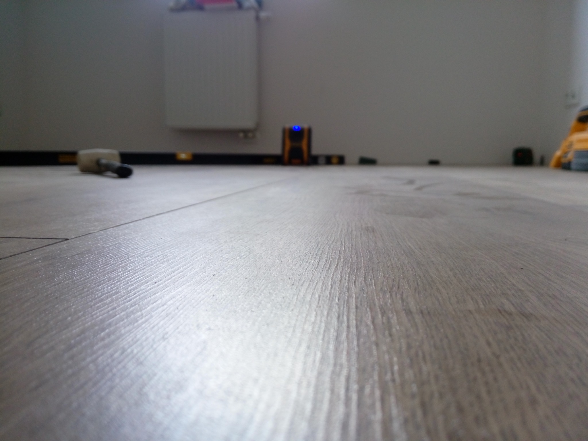 Ułożona podłoga laminowana w odcieniach szarości, widoczny poziomica laserowa, gumowy młotek i listwa przypodłogowa w tle.