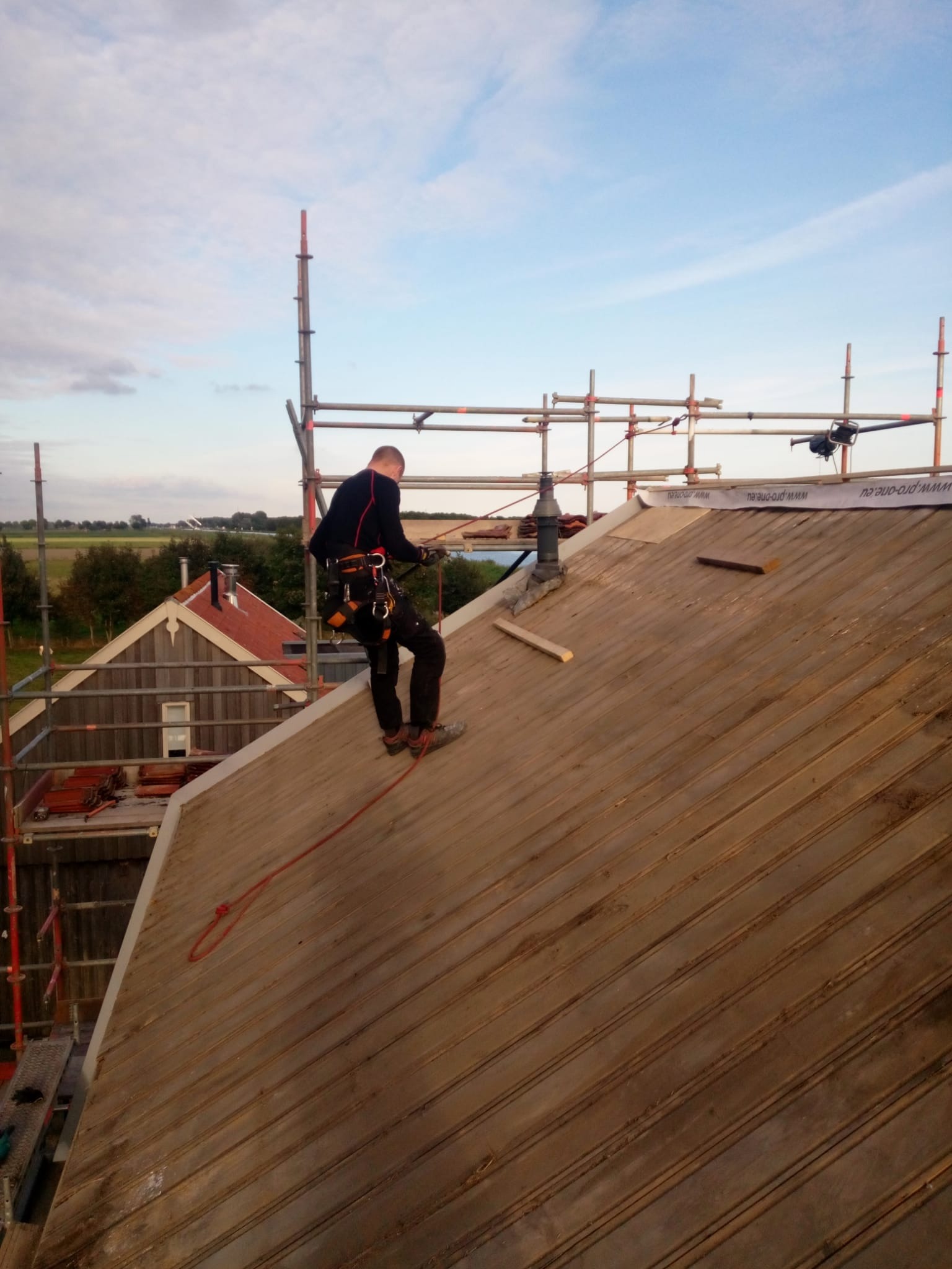 Pracownik w uprzęży zabezpieczającej stoi na drewnianym dachu podczas naprawy komina, w tle rusztowanie i zabudowania gospodarcze.