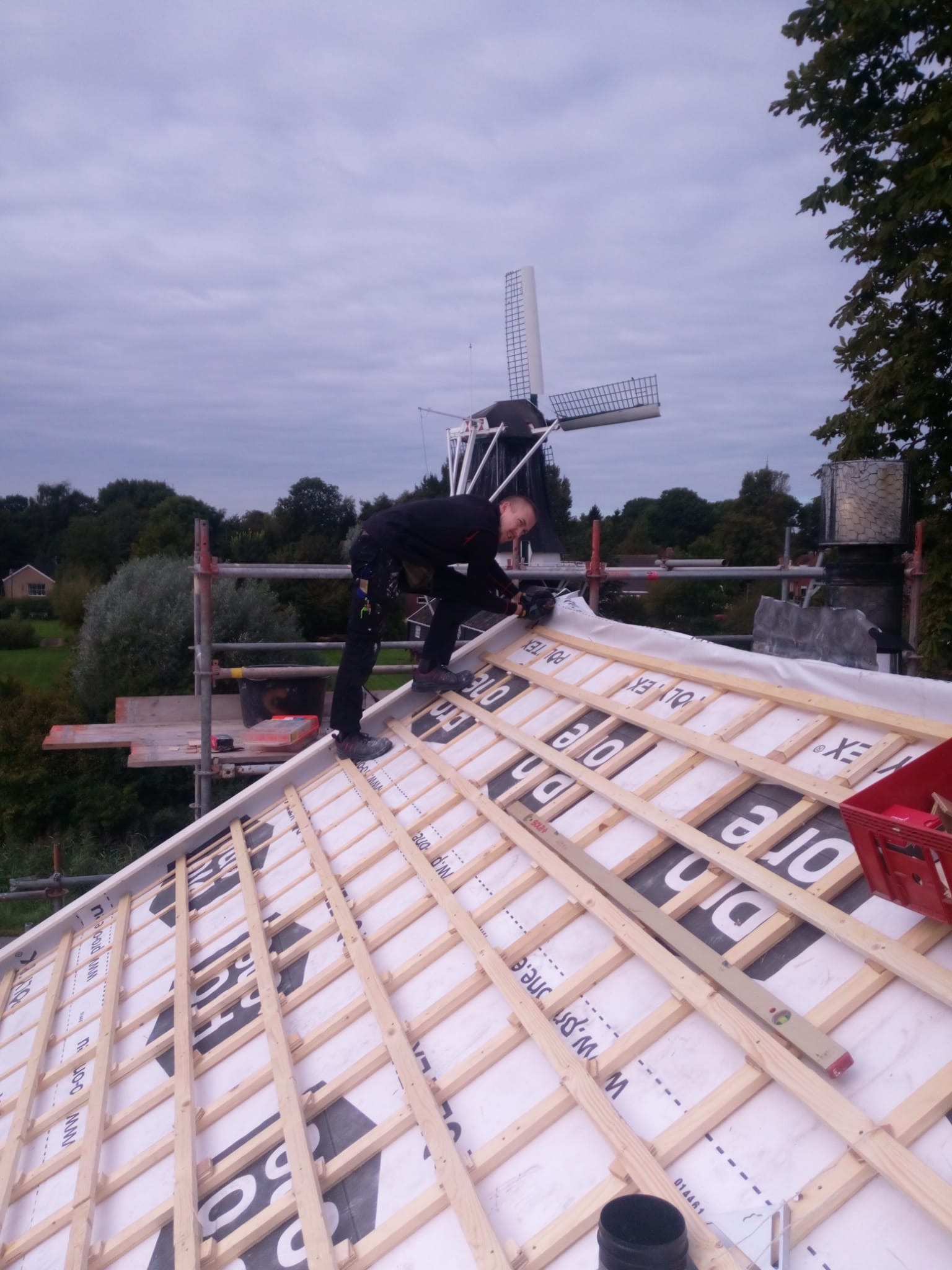 Pracownik w czarnym kombinezonie montuje drewnianą konstrukcję dachu na białej membranie, w tle widać wiatrak i drzewa.