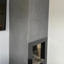 Nowoczesny kominek wykończony szarym tynkiem dekoracyjnym o fakturze betonu, z czarną obudową i półką na dekoracje, obok leży pies na posłaniu.