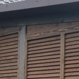 Drewniane, brązowe żaluzje zewnętrzne z widoczną fakturą drewna, zamontowane na budynku z brązowym dachem.