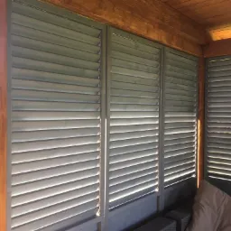 Zewnętrzne żaluzje drewniane w kolorze szarym zamontowane w drewnianej konstrukcji altany, widoczne z bliska, z delikatnym światłem słonecznym padającym na jedną z kolumn.