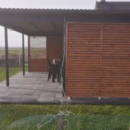 Zadaszony taras z drewnianymi panelami lamelowymi w kolorze brązowym, szarymi płytkami podłogowymi i widokiem na otwartą przestrzeń w tle.