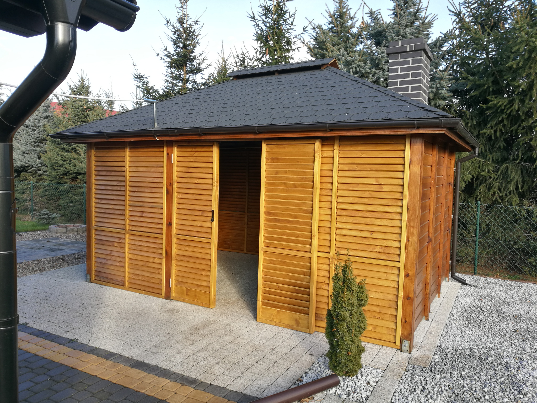 Drewniana altana z przesuwnymi, lamelowymi panelami, otwartymi na oścież, z czarnym dachem i kominem z cegły, otoczona zielenią i brukowaną nawierzchnią.