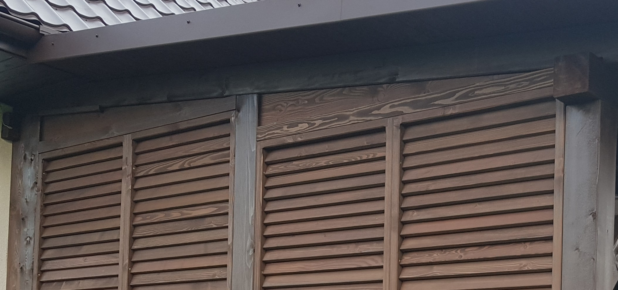 Drewniane, brązowe żaluzje zewnętrzne z widoczną fakturą drewna, zamontowane na budynku z brązowym dachem.