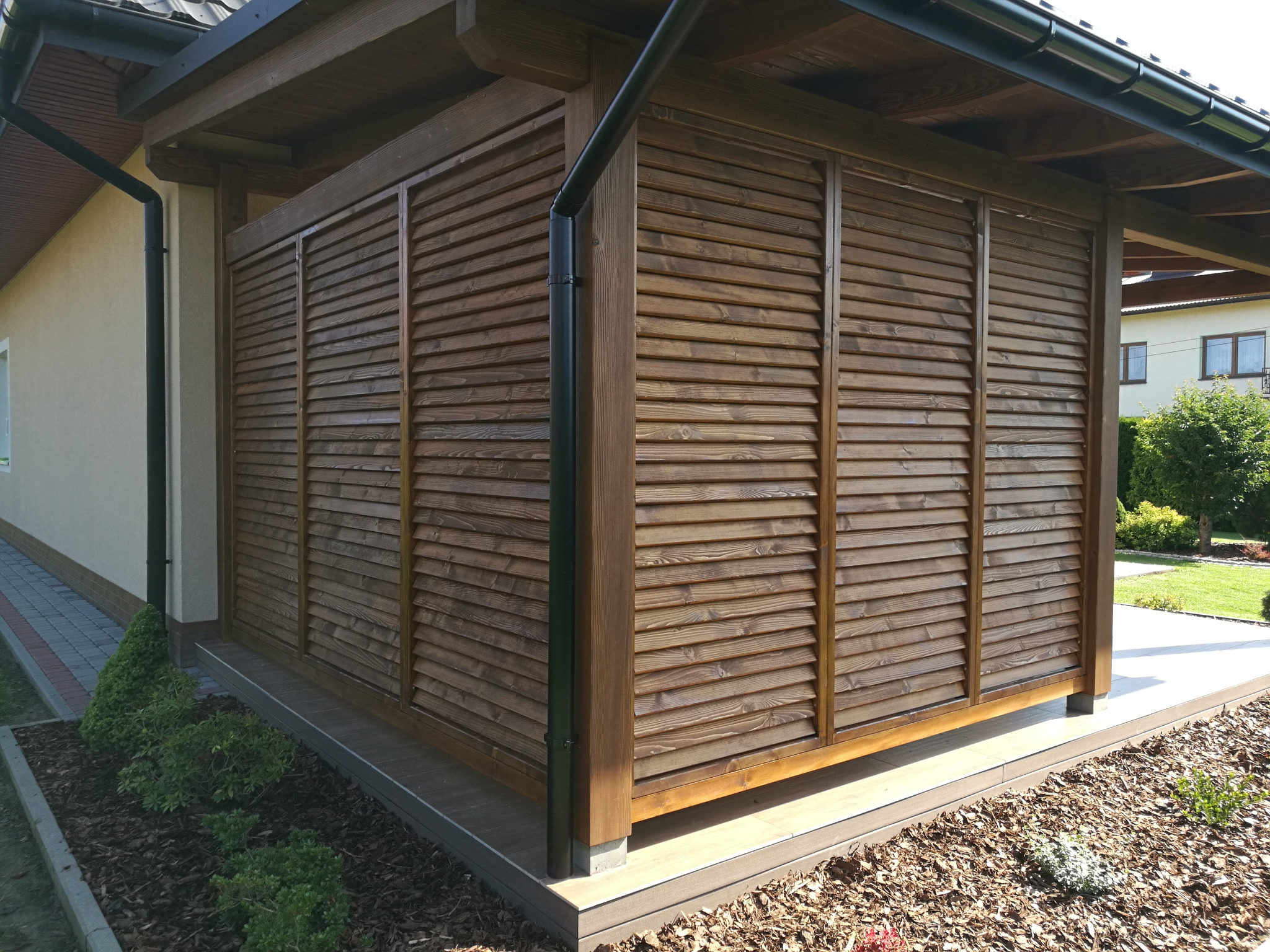 Drewniana zabudowa tarasu z żaluzjami zewnętrznymi, widok z zewnątrz, z czarną rynną i elementami architektury ogrodowej.