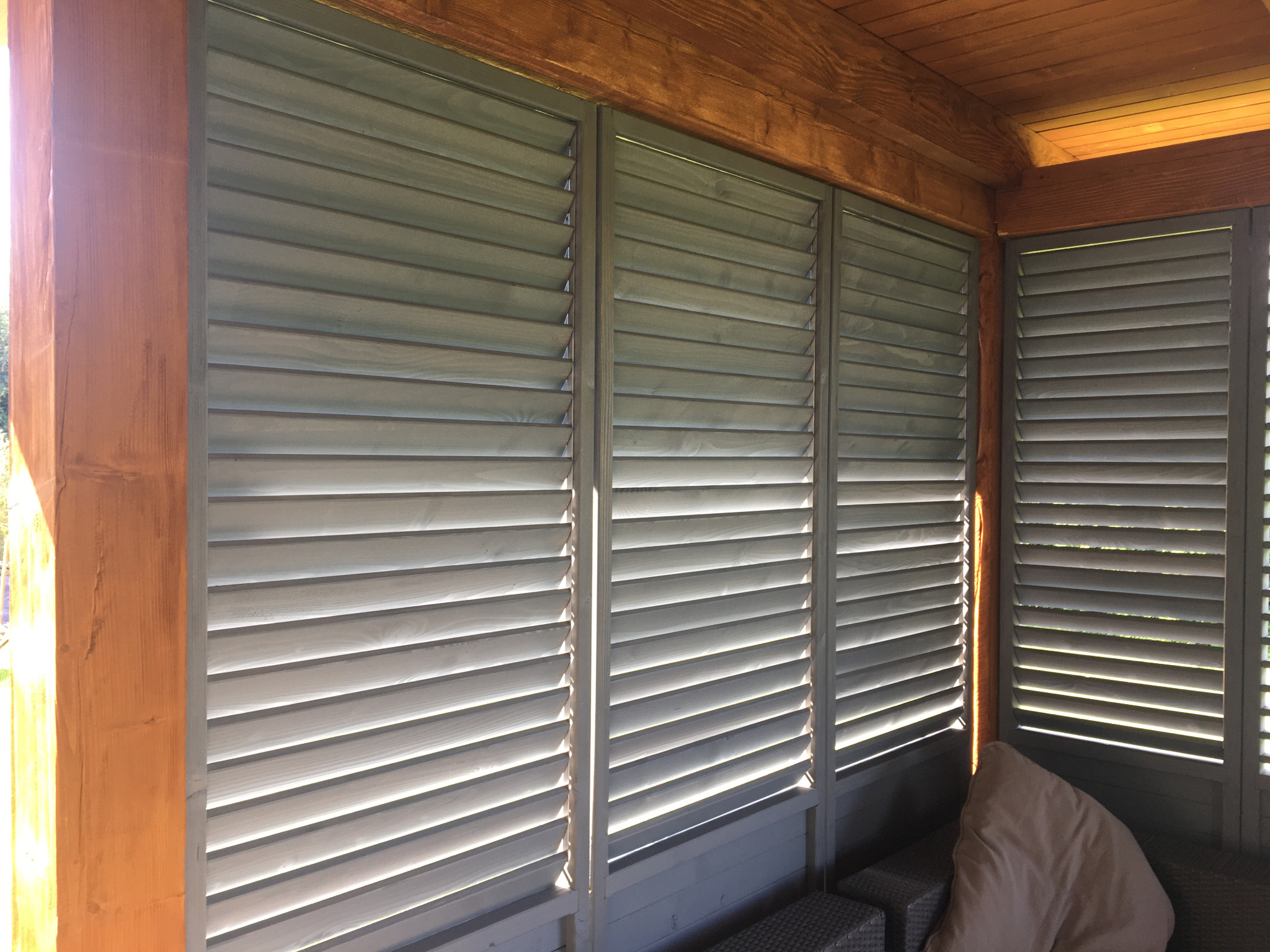 Zewnętrzne żaluzje drewniane w kolorze szarym zamontowane w drewnianej konstrukcji altany, widoczne z bliska, z delikatnym światłem słonecznym padającym na jedną z kolumn.
