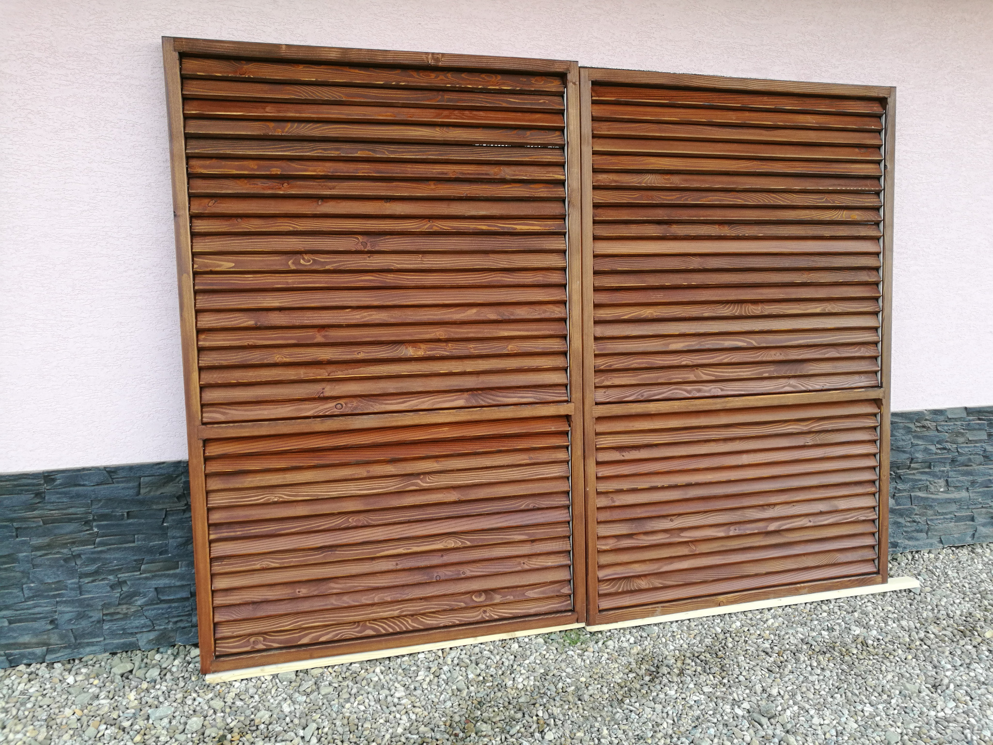 Dwa panele z ciemnego drewna z poziomymi listwami, prawdopodobnie przygotowane do montażu jako elementy osłonowe, leżące na żwirze przy ścianie budynku wykończonej kamieniem i tynkiem.