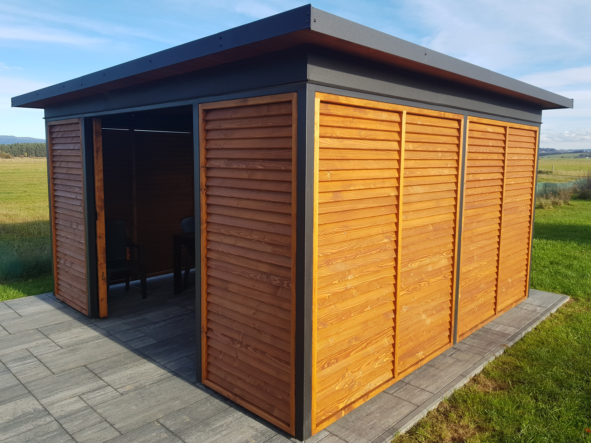 Zadaszona altana z drewnianymi żaluzjami w Wróblówce, z otwartym wejściem i widocznym wnętrzem z krzesłami, na tle zielonego trawnika i błękitnego nieba.