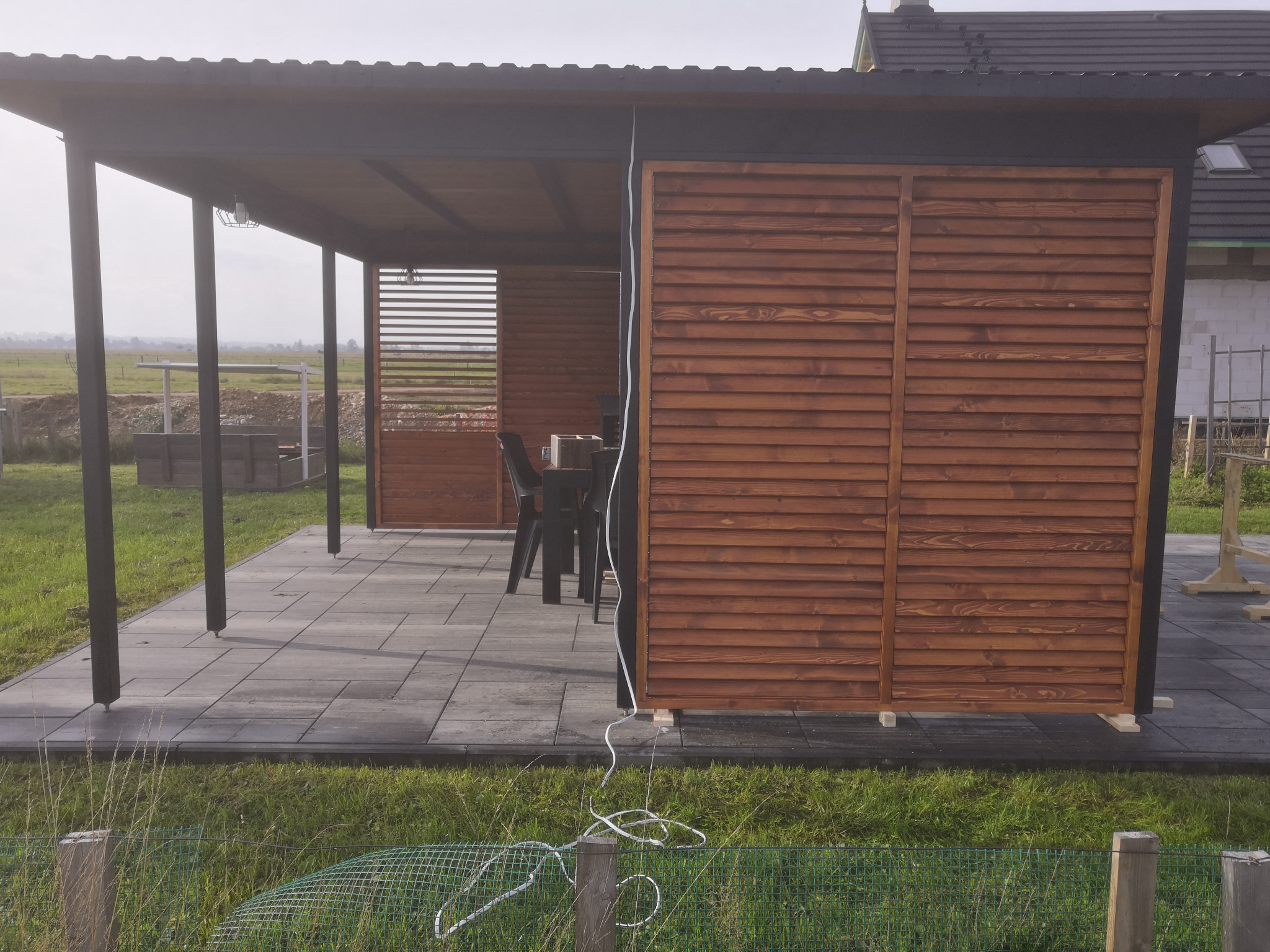 Zadaszony taras z drewnianymi panelami lamelowymi w kolorze brązowym, szarymi płytkami podłogowymi i widokiem na otwartą przestrzeń w tle.