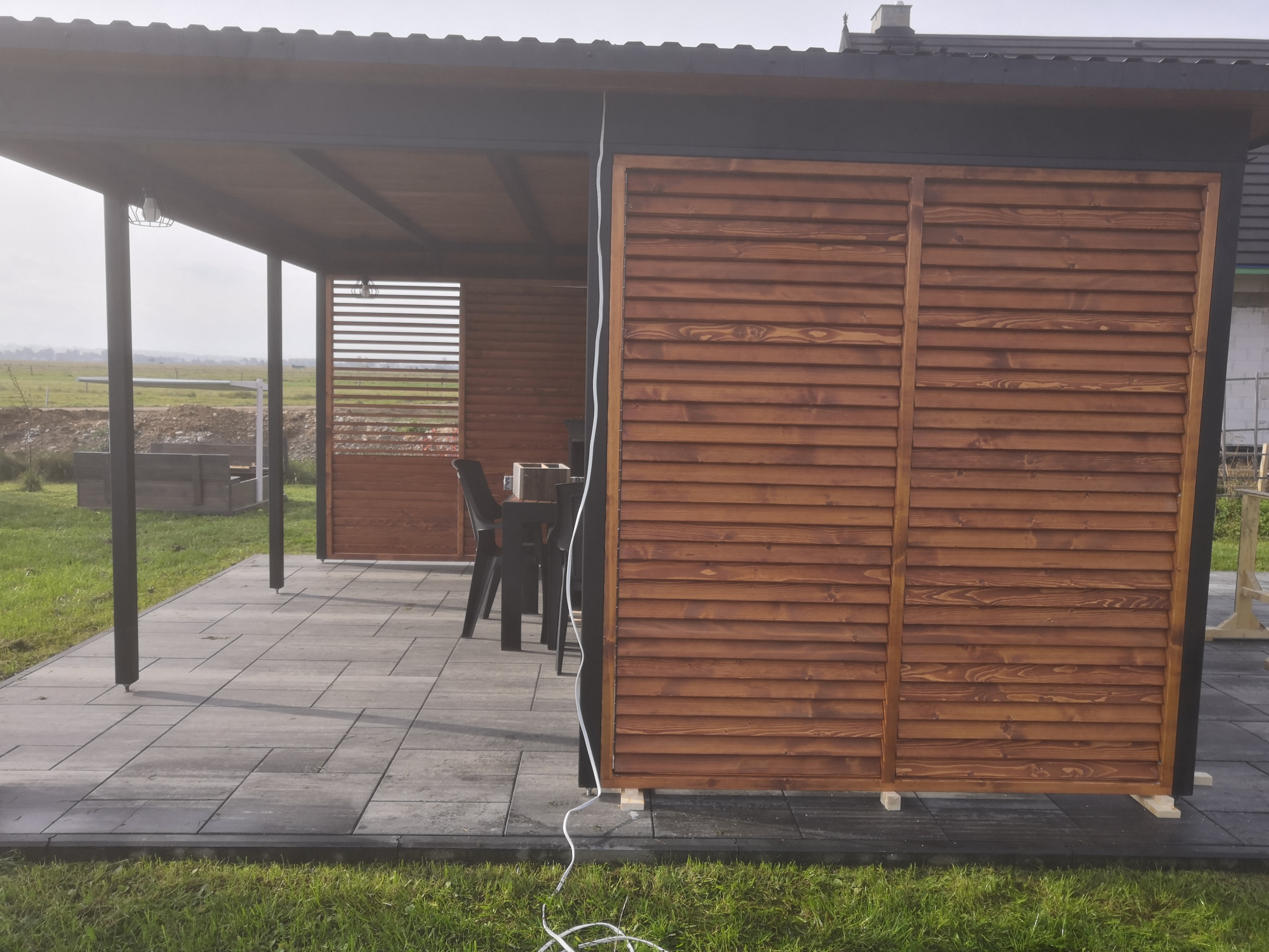 Zadaszona drewniana pergola z żaluzjowymi ścianami bocznymi, widok na polną przestrzeń w tle.