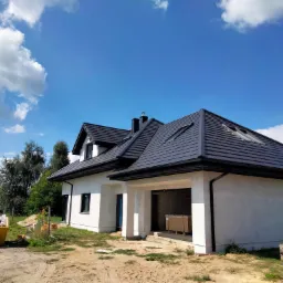 Nowoczesny dom jednorodzinny z ciemną dachówką karpiówką i oknami dachowymi, widziany z zewnątrz w słoneczny dzień, z widocznym kontenerem na śmieci budowlane i paletami w otoczeniu.