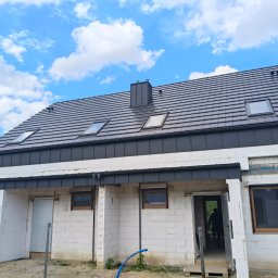 Rafał Sobkowiak - Dom w trakcie budowy z ciemnoszarym dachem z dachówką, oknami dachowymi i kominem. Ściany z białego betonu komórkowego, widoczne okna i drzwi. Błękitne niebo z chmurami w tle.