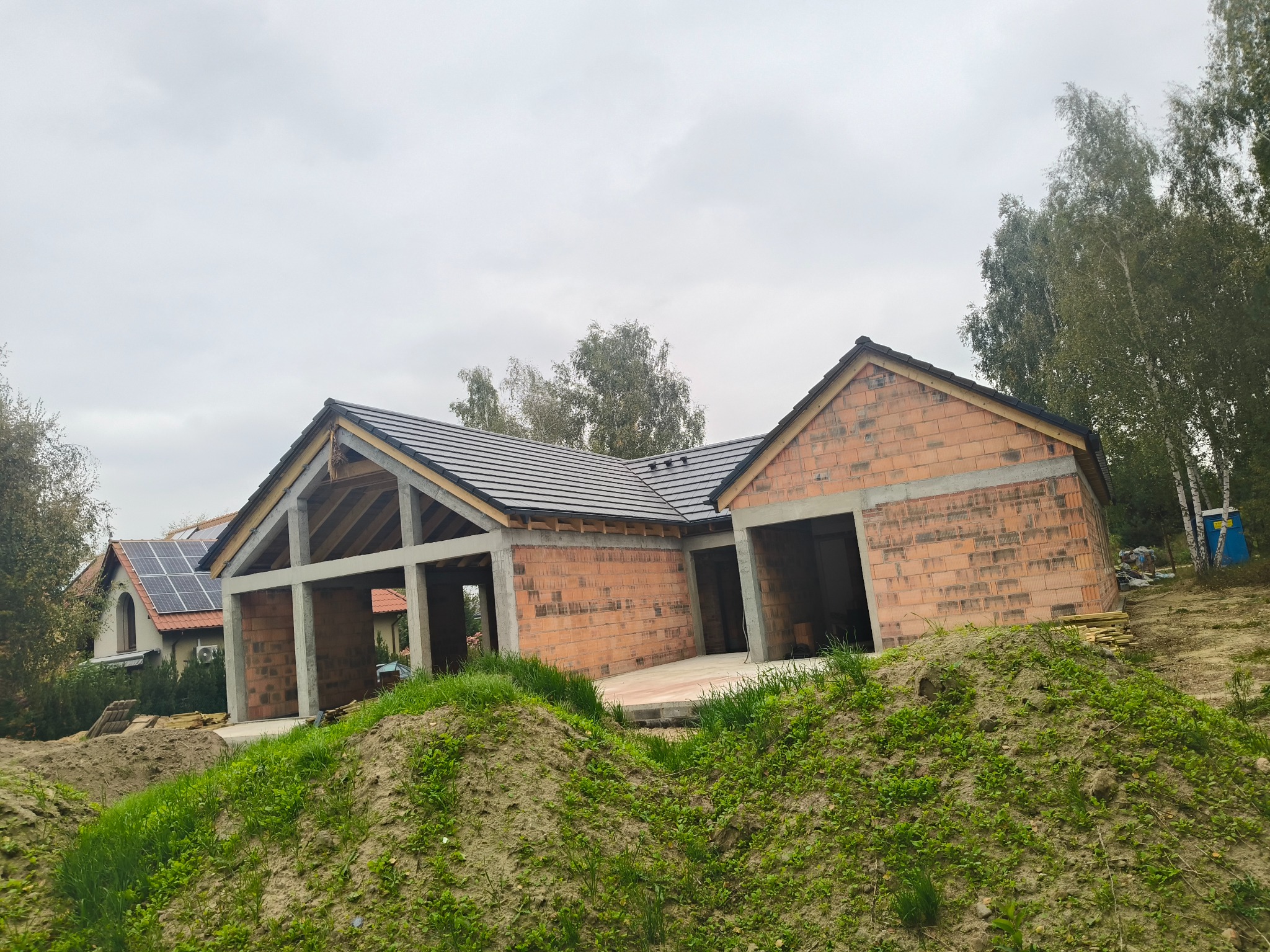 Budowa domu jednorodzinnego w stanie surowym otwartym z częściowo położonym dachem z dachówki ceramicznej. Widoczne ściany z czerwonej cegły i elementy więźby dachowej.