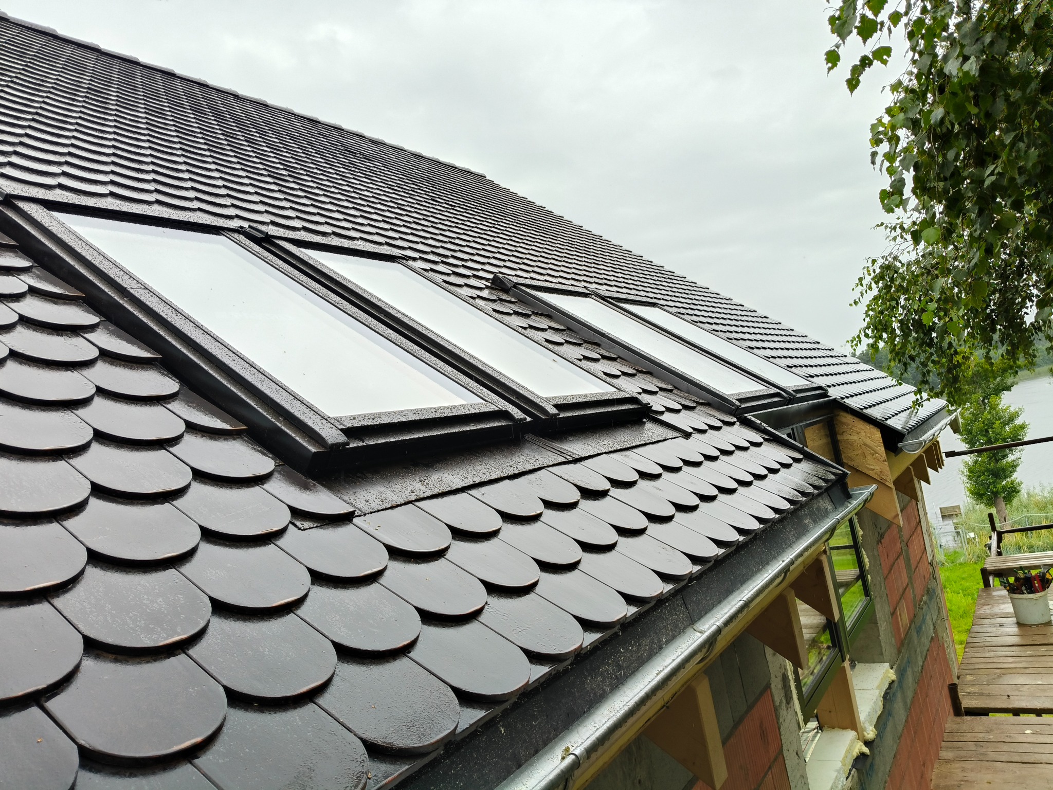 Dach pokryty ciemną dachówką bitumiczną w kształcie łuski, z trzema oknami dachowymi. Widoczny fragment rynny i elewacji w trakcie wykończenia.