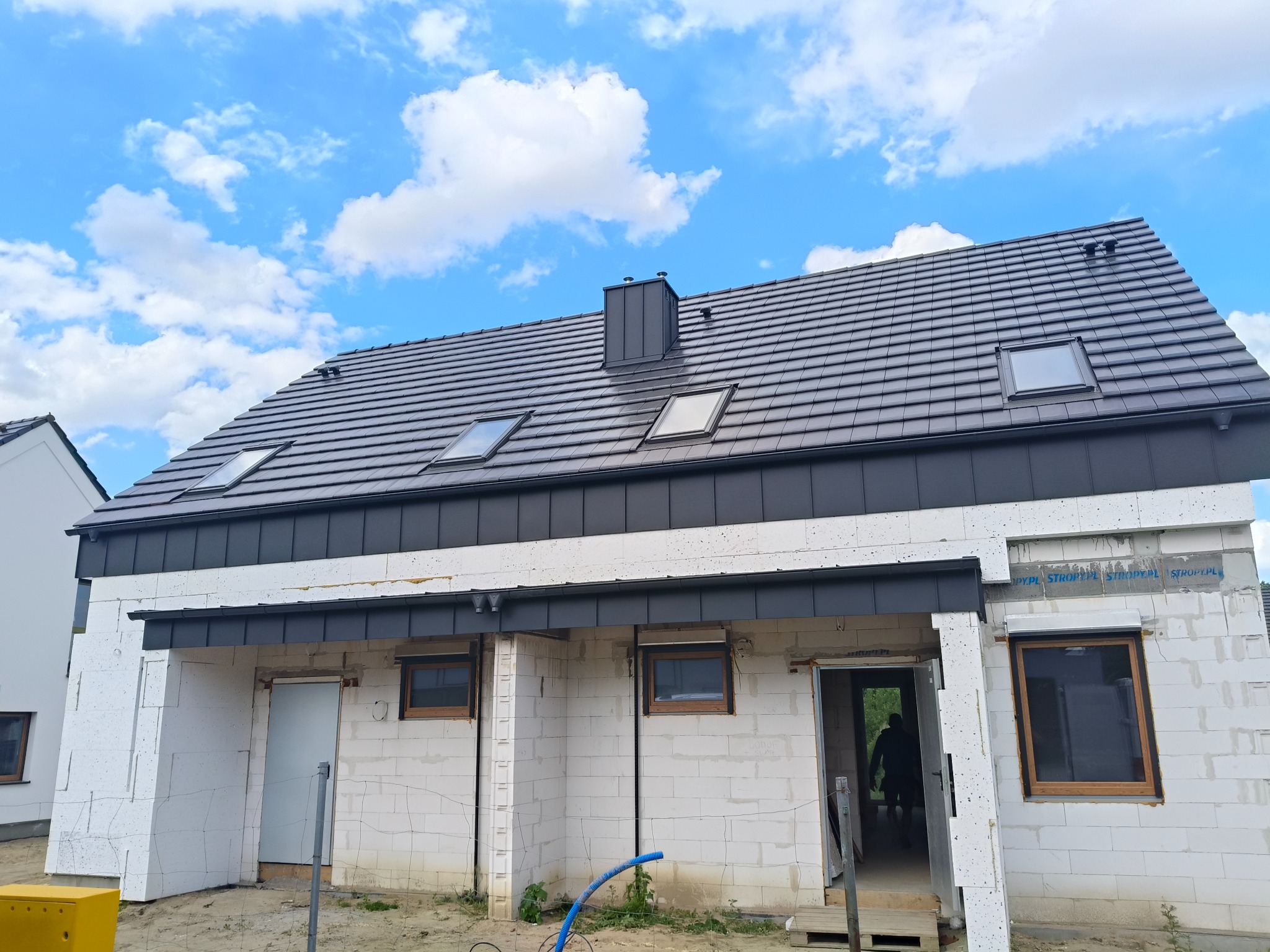 Dom w trakcie budowy z ciemnoszarym dachem z dachówką, oknami dachowymi i kominem. Ściany z białego betonu komórkowego, widoczne okna i drzwi. Błękitne niebo z chmurami w tle.