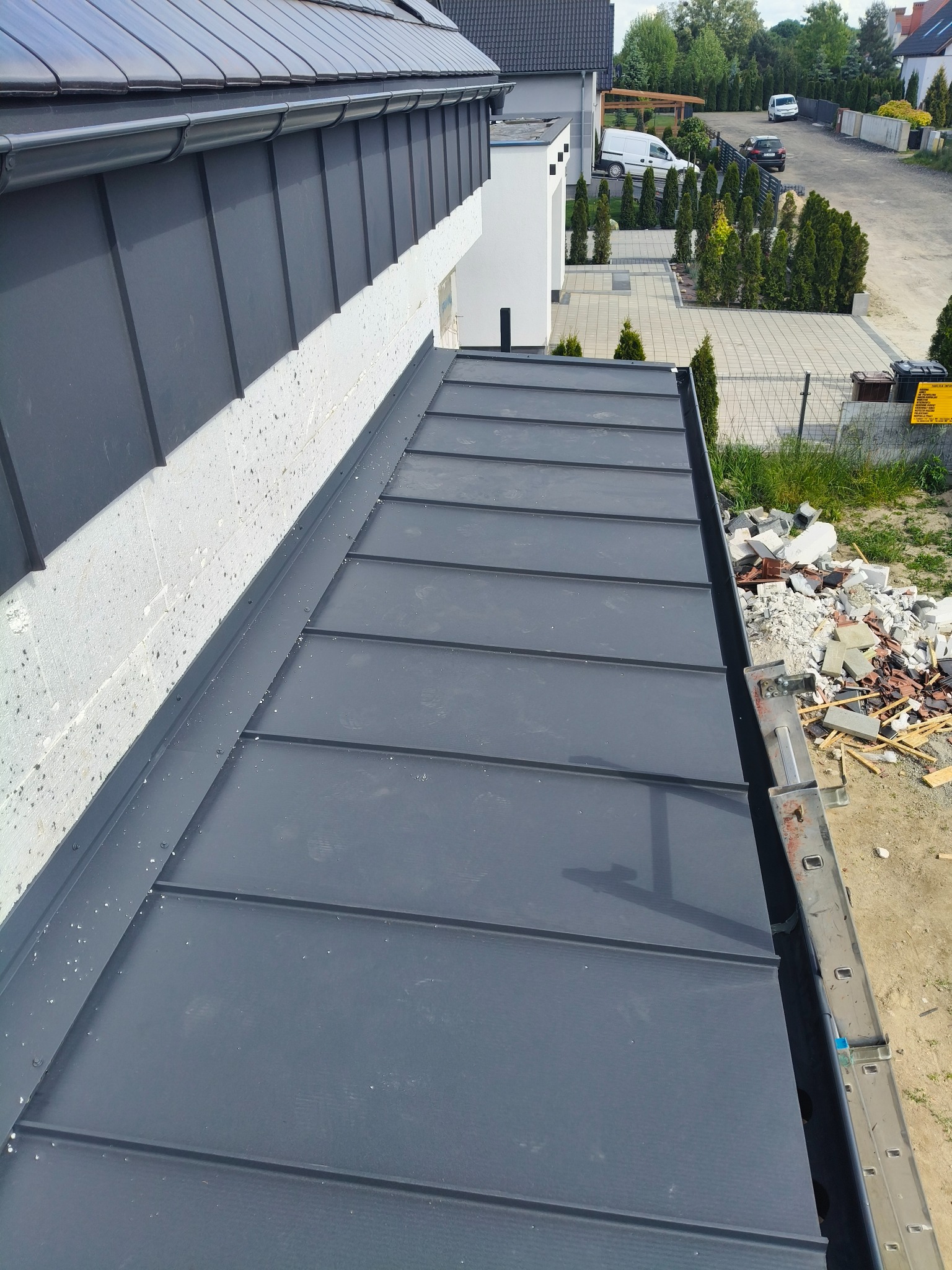 Szary dach z blachy na nowym domu jednorodzinnym, widok z góry. Fragment drabiny i gruz obok dachu. W tle zabudowa osiedlowa i zieleń.