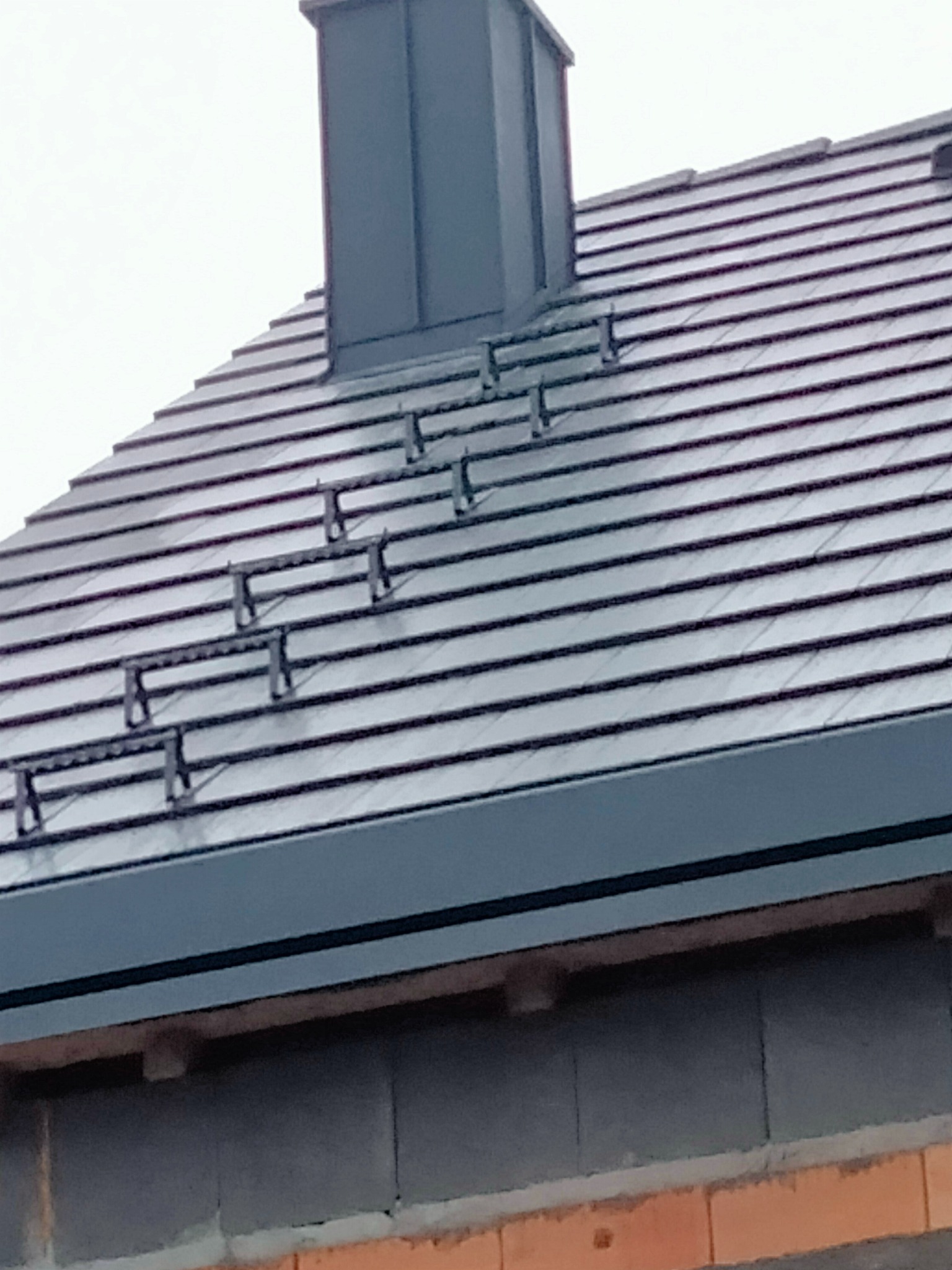 Szary dach z dachówką ceramiczną, kominem i stopniami kominiarskimi. Widok z dołu, fragment ściany budynku w tle.