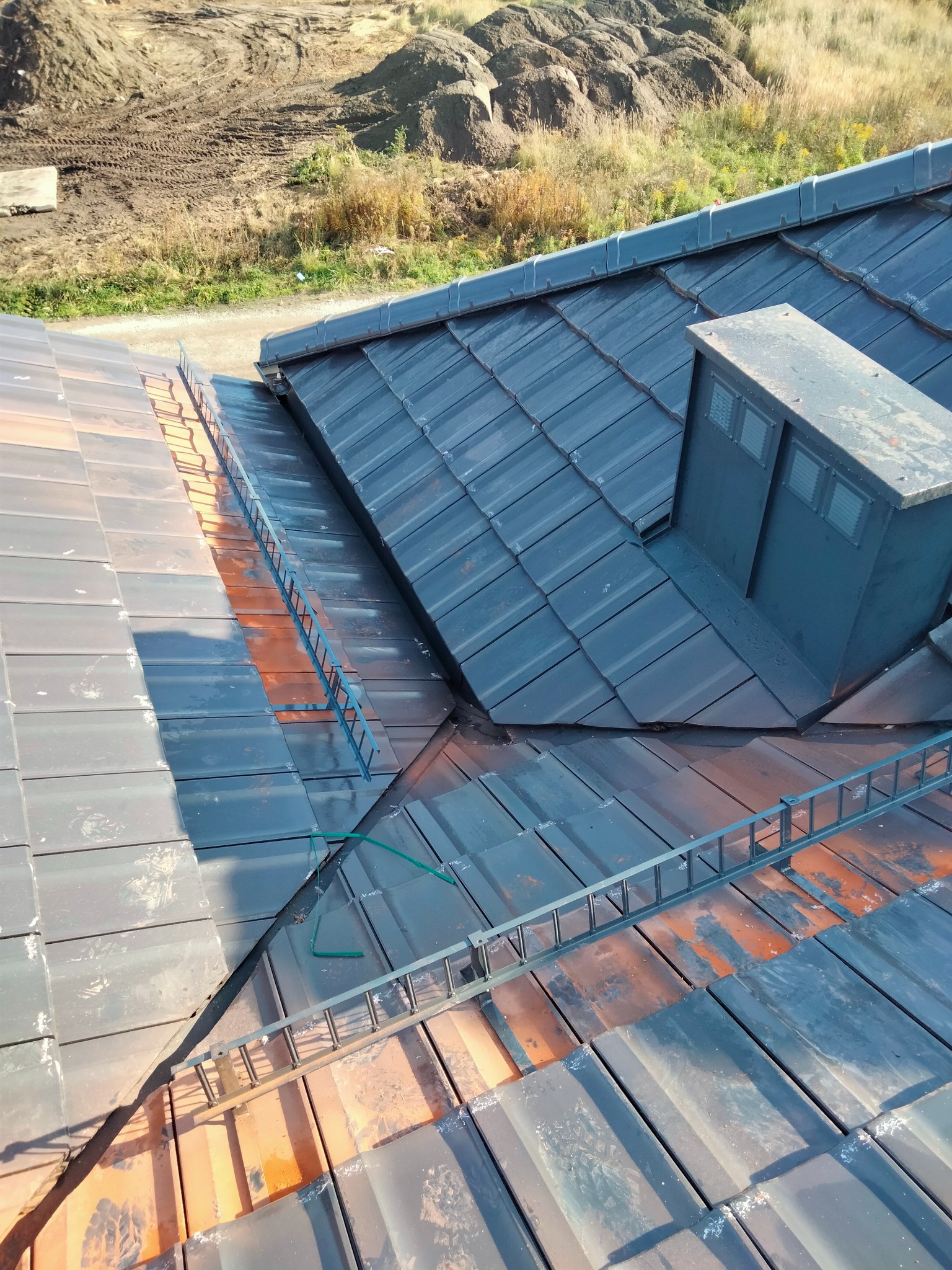 Ukończony dach z ciemnej dachówki ceramicznej, widoczny komin i zamontowane zabezpieczenia przeciwśniegowe. W tle ziemia i roślinność.